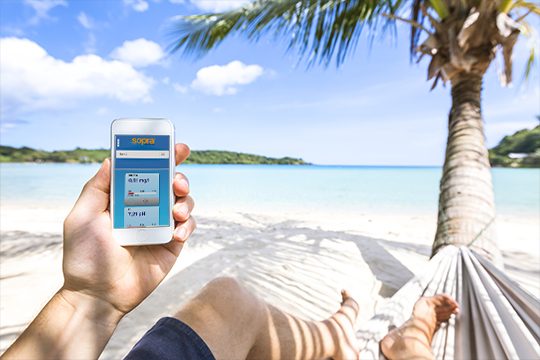 Ihren Smart Pool behalten Sie auch während des Strandurlaubs im Blick. Jemand liegt in einer Hängematte mit Meerblick und kontrolliert die Wasserwerte seines Smart Pools per Handy.