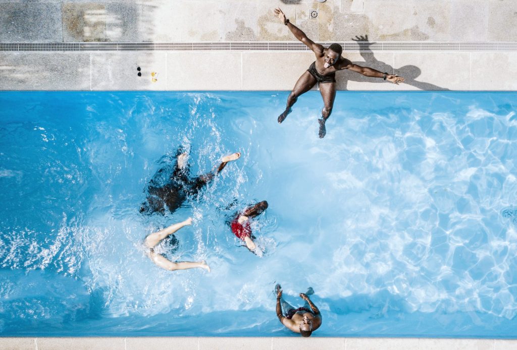 Aufnahme einer Gruppe aus mehreren Personen aus der Vogelperspektive. Sie sitzen am Pool oder schwimmen darin und sind fröhlich.