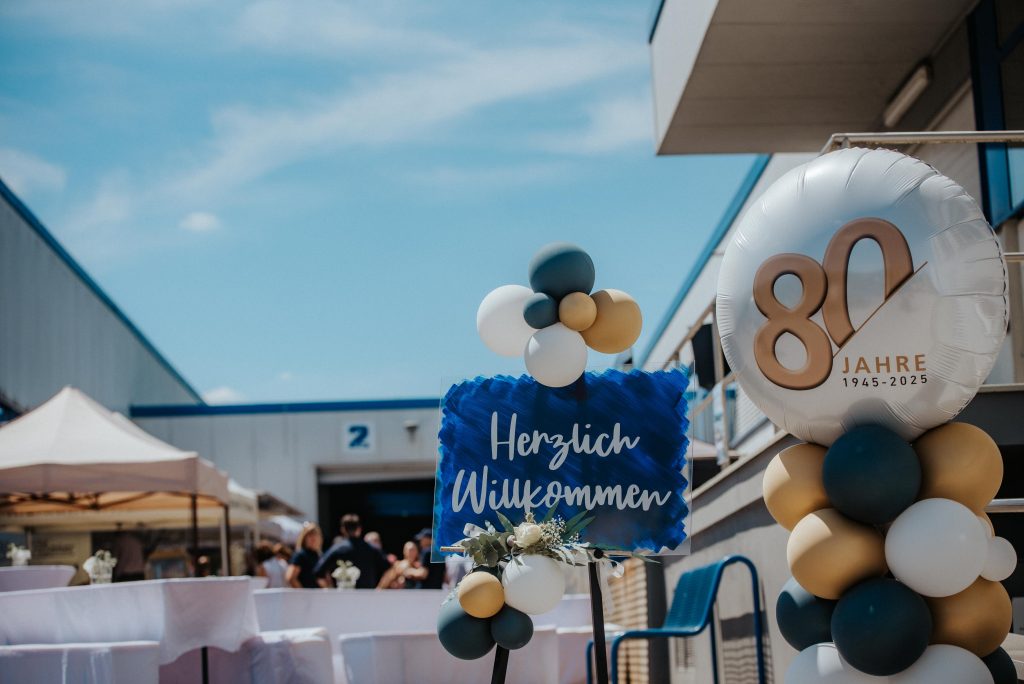Ein Schild auf dem Herzlich Willkommen steht, wird von Luftballons umrahmt. daneben befindet sich ein Luftballon mit einer großen 80.