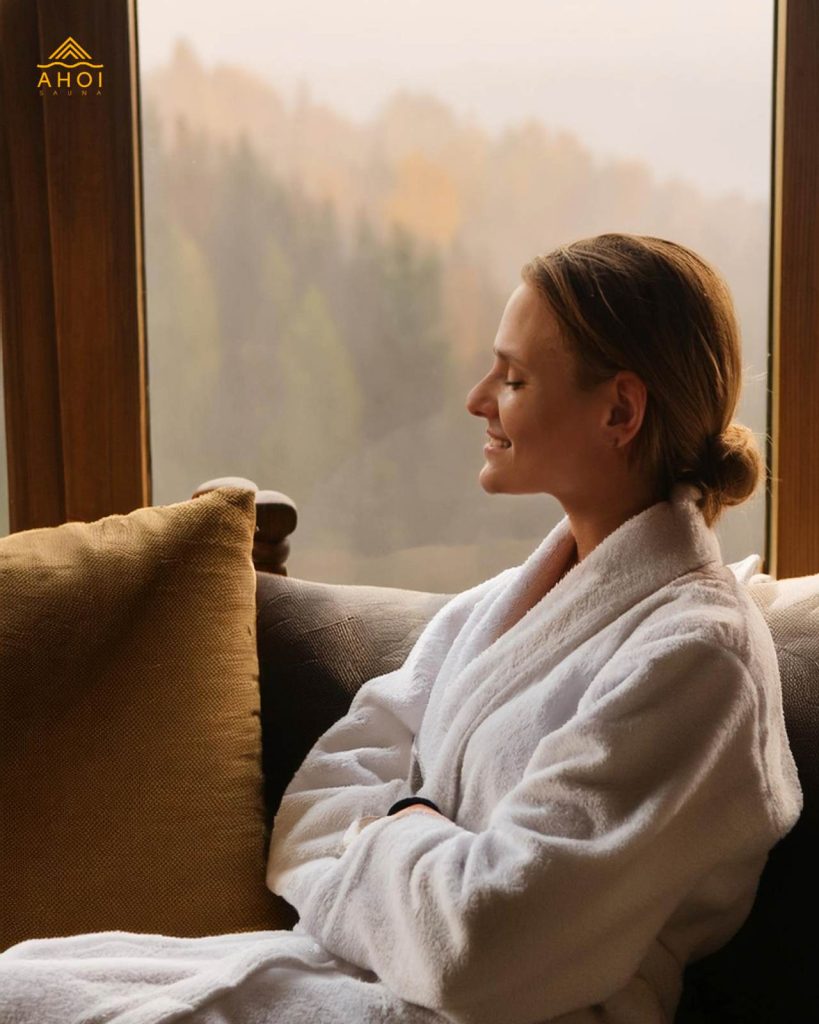 Eine Frau im Bademantel sitzt entspannt vor einem Fenster. Sie hat die Augen geschlossen.